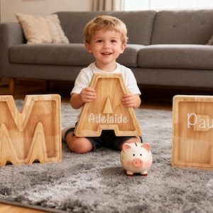 May include: A child sits with wooden letter-shaped piggy banks spelling out names, and a small ceramic piggy bank. The wooden letters are light brown with names engraved in white. The piggy bank is pink and white.