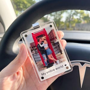 Peut inclure: Porte-clés en acrylique transparent avec une photo d'un couple s'embrassant devant une cabine téléphonique rouge. Le porte-clés a un clip argenté et les mots "My entire world" imprimés en bas.