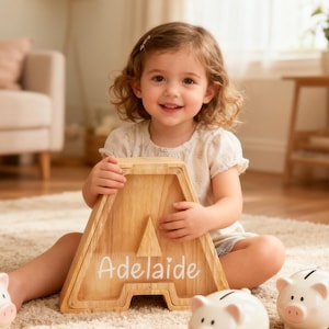Op de afbeelding: Een kind houdt een houten letter A spaarpot vast met de naam "Adelaide" in witte letters. De spaarpot is lichtbruin met een natuurlijke houtnerf. Twee witte spaarvarkens staan op de voorgrond.