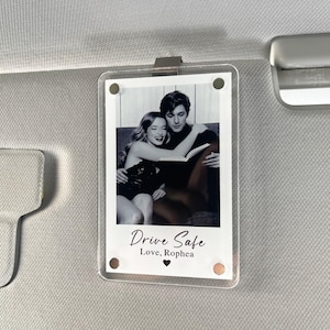 May include: A clear acrylic photo holder featuring a monochrome image of a couple. The text "Drive Safe, Love, Rophea" is printed below the photo. The holder is attached to a car sun visor with a silver clip.