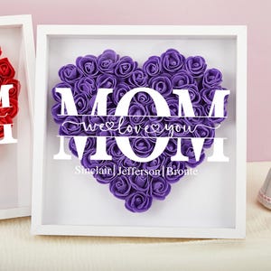 Puede incluir: Marco blanco con un corazón morado hecho de rosas de espuma. El corazón tiene la palabra "MOM" escrita en letras blancas con el texto "we love you" en el medio. El nombre "Sinclair Jefferson Bronte" está escrito debajo del corazón.