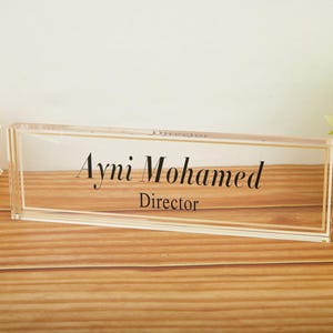 Può includere: Un'elegante targhetta da scrivania in acrilico trasparente con bordi dorati, che mostra il nome "Ayni Mohamed" e il titolo "Director" in elegante scrittura nera. La targhetta poggia su una superficie di legno, con accenti floreali sullo sfondo.