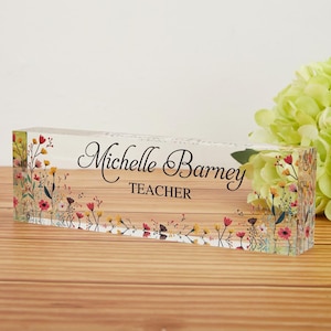 May include: A clear acrylic desk block with the name "Michelle Barney" and the title "TEACHER" in black script. The block is decorated with colorful floral designs and rests on a wooden surface.