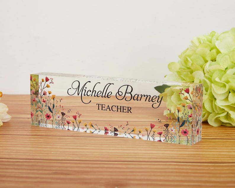 May include: A clear acrylic desk block with the name "Michelle Barney" and the title "TEACHER" in black script. The block is decorated with colorful floral designs and rests on a wooden surface.