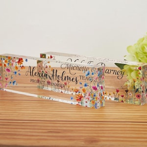 May include: Two clear acrylic desk nameplates with floral designs. One plate reads "Alexia Holmes, Project Manager" in black script. The other plate has a name and the title "Teacher". The plates are decorated with colorful flowers.
