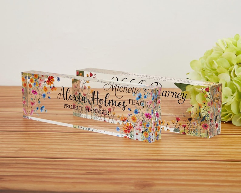 May include: Two clear acrylic desk nameplates with floral designs. One plate reads "Alexia Holmes, Project Manager" in black script. The other plate has a name and the title "Teacher". The plates are decorated with colorful flowers.