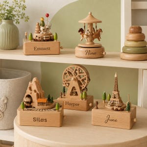 May include: Five wooden music boxes with personalized names Emmett, Nina, Silas, Harper, and June. The designs include a train with bears, a carousel with horses, a mountain tunnel with a train, a Ferris wheel, and the Eiffel Tower with trees.