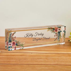 May include: A clear acrylic desk nameplate with floral accents and the text "Kelly Farley Digital Fraud." The nameplate has a gold border and is decorated with pink and white roses and greenery.