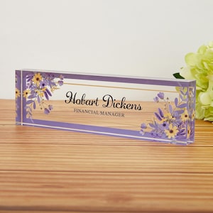May include: A rectangular desk nameplate with the name "Hobart Dickens" and the title "Financial Manager" in elegant script. The clear acrylic block features a lavender border and floral accents in yellow and purple.