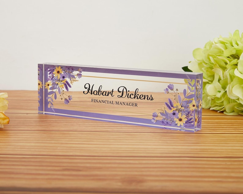 May include: A rectangular desk nameplate with the name "Hobart Dickens" and the title "Financial Manager" in elegant script. The clear acrylic block features a lavender border and floral accents in yellow and purple.