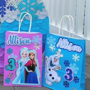 Puede incluir: Dos bolsas de regalo azules y rosas con asas blancas. Las bolsas est&aacute;n decoradas con personajes de Frozen, copos de nieve, el nombre "Allison" y el n&uacute;mero "3".