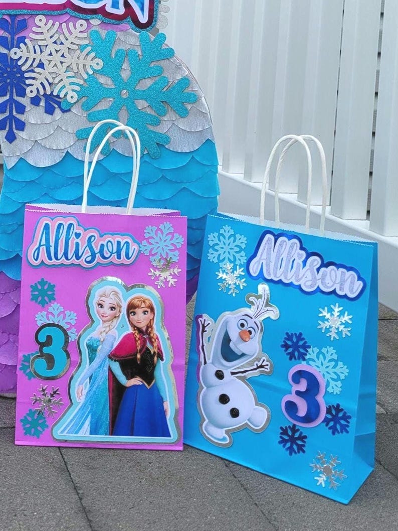 Puede incluir: Dos bolsas de regalo azules y rosas con asas blancas. Las bolsas est&aacute;n decoradas con personajes de Frozen, copos de nieve, el nombre "Allison" y el n&uacute;mero "3".