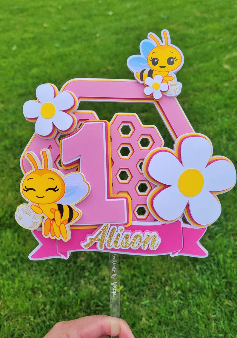 以下が含まれることがあります： ピンクと黄色のケーキトッパー。数字の1、花、漫画のミツバチが描かれています。「Alison」という名前が金色の文字で書かれています。1歳の誕生日のお祝いに最適です。