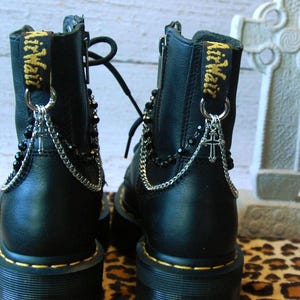 Op de afbeelding: Zwarte leren laarzen met zilveren kettingen en een zilveren kruisbedel. De laarzen hebben een geel label met de tekst "Dr. Martens".