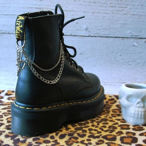 May include: Black leather platform boot with silver chain and star accents. The boot has yellow stitching and a chunky sole. A small white skull-shaped ceramic container sits next to the boot on a leopard print surface.