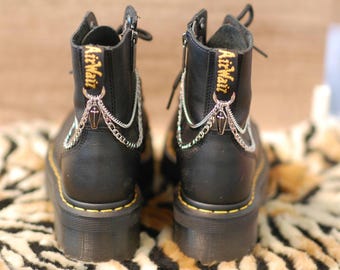 Puede incluir: Botas de cuero negro con detalles de costura amarilla y cadenas y abalorios plateados. Las botas tienen una cremallera en la parte posterior y el texto "AirWair" es visible en la parte posterior de las botas.