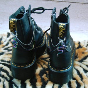 Puede incluir: Botas de cuero negro con suela gruesa y una cadena plateada con un dije de serpiente que cuelga de cada bota. Las botas tienen el texto "AirWair" bordado en el lateral.