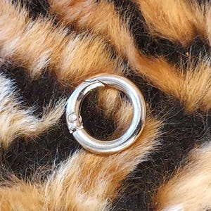 May include: A silver-colored, circular clasp with a hinged opening. The clasp is set against a backdrop of faux fur in shades of brown, black, and cream, resembling a leopard print.