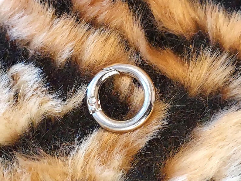 May include: A silver-colored, circular clasp with a hinged opening. The clasp is set against a backdrop of faux fur in shades of brown, black, and cream, resembling a leopard print.