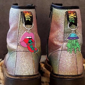Könnte beinhalten: Ein Paar schwarze Glitzer-Stiefel mit buntem Regenbogen-Glitter. Die Stiefel haben einen schwarzen Schnürverschluss und eine schwarze Gummisohle. An jedem Stiefel ist ein Anhänger an den Schnürsenkeln befestigt. Der Anhänger am linken Stiefel ist eine rosa Zunge mit einem grünen Außerirdischen darauf. Der Anhänger am rechten Stiefel ist ein grünes außerirdisches Raumschiff mit dem Text "Get in Loser."