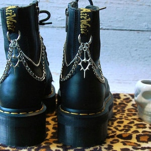 Puede incluir: Botas negras de plataforma con detalles de cadenas y abalorios plateados. Las botas tienen una suela cosida amarilla y cierre de cremallera. Un recipiente de cerámica con forma de calavera está cerca. Las botas están sobre una superficie con estampado de leopardo.