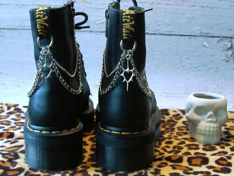May include: Black platform boots with silver chain and charm accents. The boots have a yellow stitched sole and a zipper closure. A skull-shaped ceramic container sits nearby. The boots are on a leopard print surface.