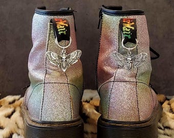 May include: A pair of black glitter Doc Martens boots with silver moth charms attached to the laces. The boots have a rainbow iridescent glitter finish and the laces have a black tag that says "Dr. Martens".
