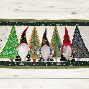 May include: A green, white and red Christmas-themed quilted wall hanging with five gnomes standing in front of five Christmas trees. The gnomes are wearing red, blue and green hats. The trees are decorated with gold and silver baubles. The text "Merry Christmas" is written on the bottom of the wall hanging.