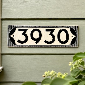 May include: A rectangular house number plaque featuring the digits 3930 in bold black font on a cream-colored background. The plaque is framed by a black border with vertical line detailing and decorative black leaf-like shapes at each end.