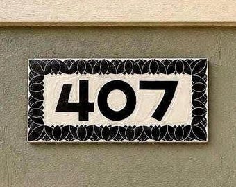 May include: A rectangular house number plaque displaying the digits 407 in bold black font against a cream-colored background, framed by a decorative black border featuring a repeating leaf or petal pattern mounted on a porch pillar.