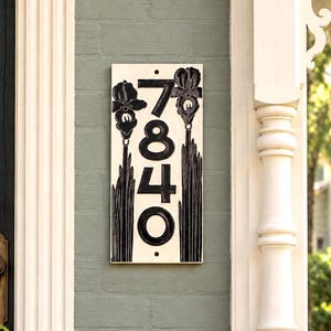 May include: A rectangular ceramic house number plaque featuring the digits 7840 in a bold black font. The plaque has a cream-colored background and is decorated with black floral illustrations of irises on the left and right sides.