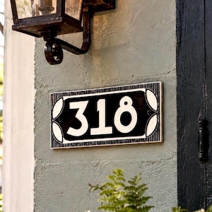 May include: A rectangular house number plaque featuring the white digits 318 centered on a black background with a white border. The plaque has decorative white leaf-like accents at the corners and is mounted on a light gray textured wall.