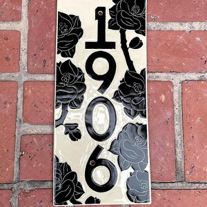 May include: A rectangular ceramic house number plaque with a cream-colored background and black floral illustrations. The vertical numbers 1906 are printed in black in the center, with two circular mounting holes positioned above and below the numbers.