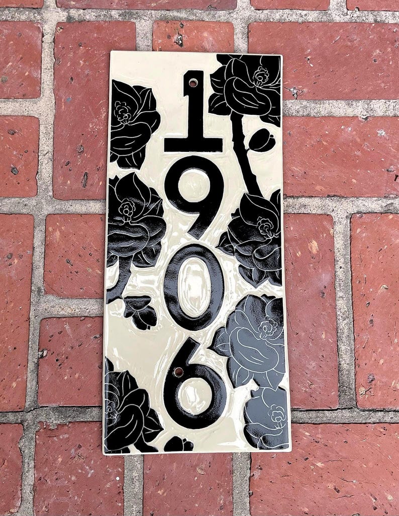 May include: A rectangular ceramic house number plaque with a cream-colored background and black floral illustrations. The vertical numbers 1906 are printed in black in the center, with two circular mounting holes positioned above and below the numbers.