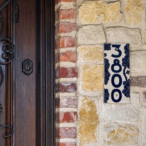 May include: A rectangular ceramic house number plaque featuring the digits 3800 in a dark blue font. The plaque has a cream-colored background decorated with a dark blue floral pattern and is mounted on a stone wall next to a dark wood door.