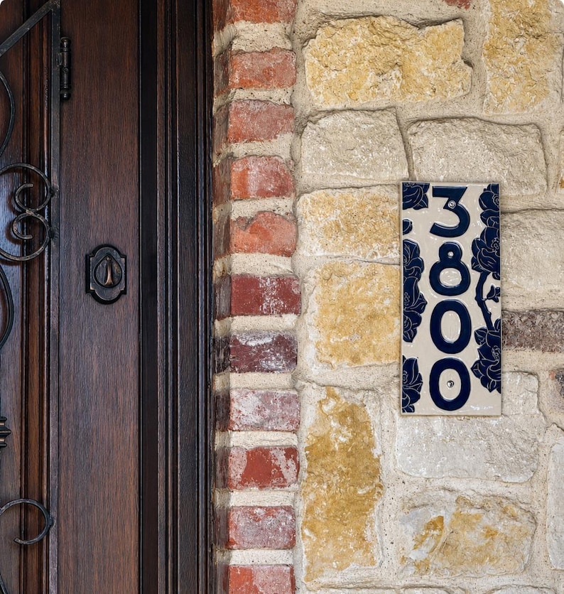 May include: A rectangular ceramic house number plaque featuring the digits 3800 in a dark blue font. The plaque has a cream-colored background decorated with a dark blue floral pattern and is mounted on a stone wall next to a dark wood door.