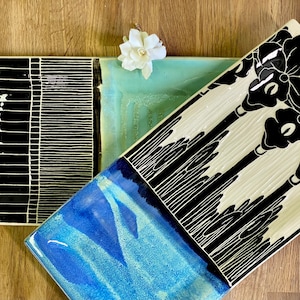 May include: Three rectangular ceramic serving trays with black and white designs. The first tray has a black background with white geometric patterns. The second tray has a light blue background with white and black abstract designs. The third tray has a white background with black floral designs.