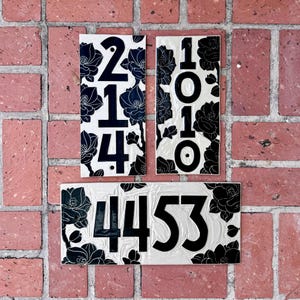 May include: Three rectangular house number plaques featuring a cream-colored background with black floral patterns. Two vertical plaques display the numbers 214 and 1010, while one horizontal plaque displays the number 4453 in bold black lettering.