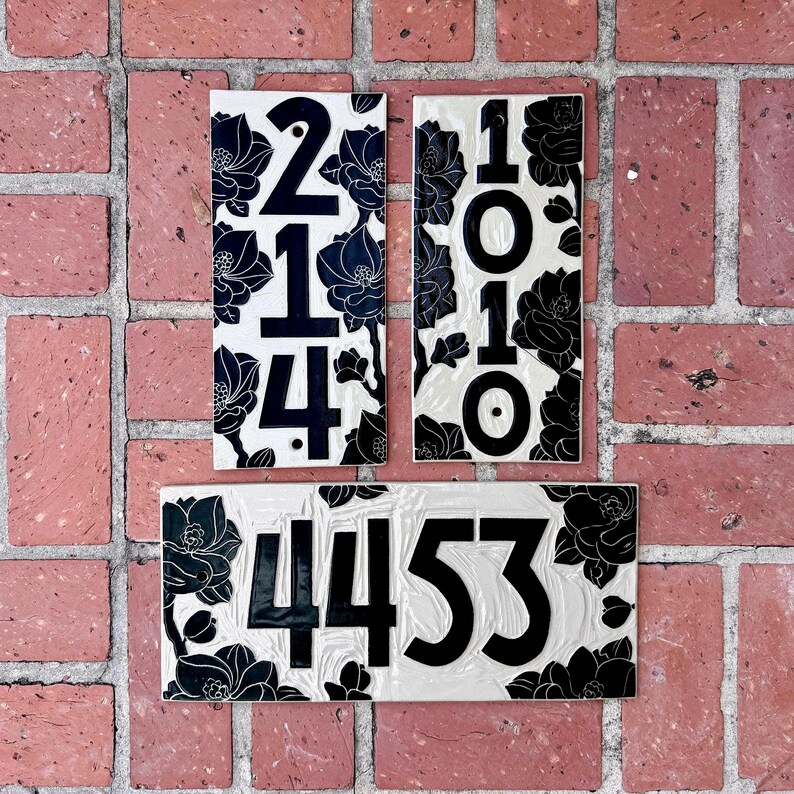 May include: Three rectangular house number plaques featuring a cream-colored background with black floral patterns. Two vertical plaques display the numbers 214 and 1010, while one horizontal plaque displays the number 4453 in bold black lettering.