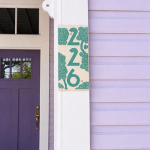 May include: A rectangular house number plaque featuring the digits 226 in a dark teal color. The plaque has a light beige background decorated with embossed dark teal floral patterns surrounding the numbers, mounted on a white wooden post.