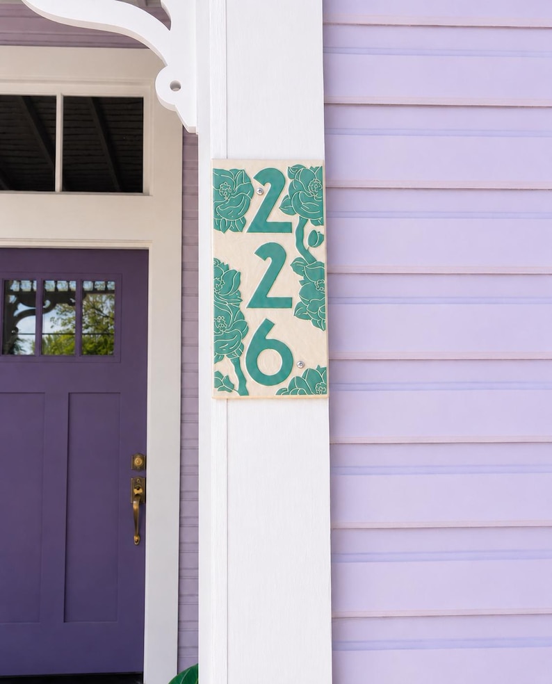 May include: A rectangular house number plaque featuring the digits 226 in a dark teal color. The plaque has a light beige background decorated with embossed dark teal floral patterns surrounding the numbers, mounted on a white wooden post.
