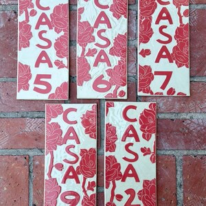May include: Five rectangular ceramic tiles featuring a cream-colored background with embossed red floral patterns and the text CASA 5, CASA 6, CASA 7, CASA 9, and CASA 2. Each tile displays a unique arrangement of red flowers and bold red lettering.