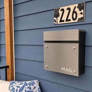 May include: A rectangular house number plaque displaying the digits 226 with black floral illustrations on a cream background, positioned above a brushed metal mailbox with a black frame and the word MAIL printed in white on the front panel.