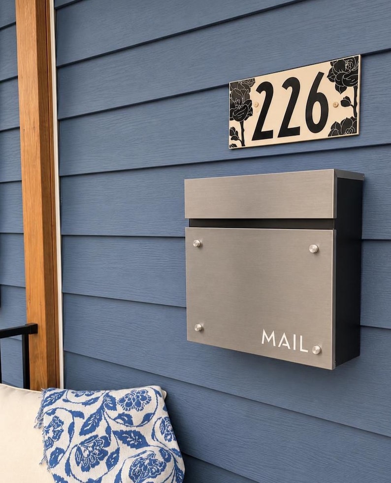 May include: A rectangular house number plaque displaying the digits 226 with black floral illustrations on a cream background, positioned above a brushed metal mailbox with a black frame and the word MAIL printed in white on the front panel.
