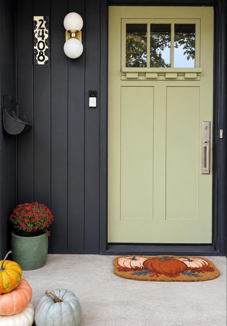 May include: A sage green front door with a rectangular window pane, set against dark charcoal vertical siding. Features include a brass handle, a double globe wall light, a house number plaque reading 2408, a pumpkin-themed doormat, and pumpkins.