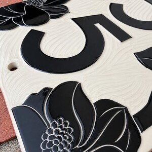 May include: A rectangular ceramic house number plaque featuring a cream-colored background with a textured, wavy surface. The plaque displays a large black numeral five and is decorated with glossy black floral motifs at the corners.