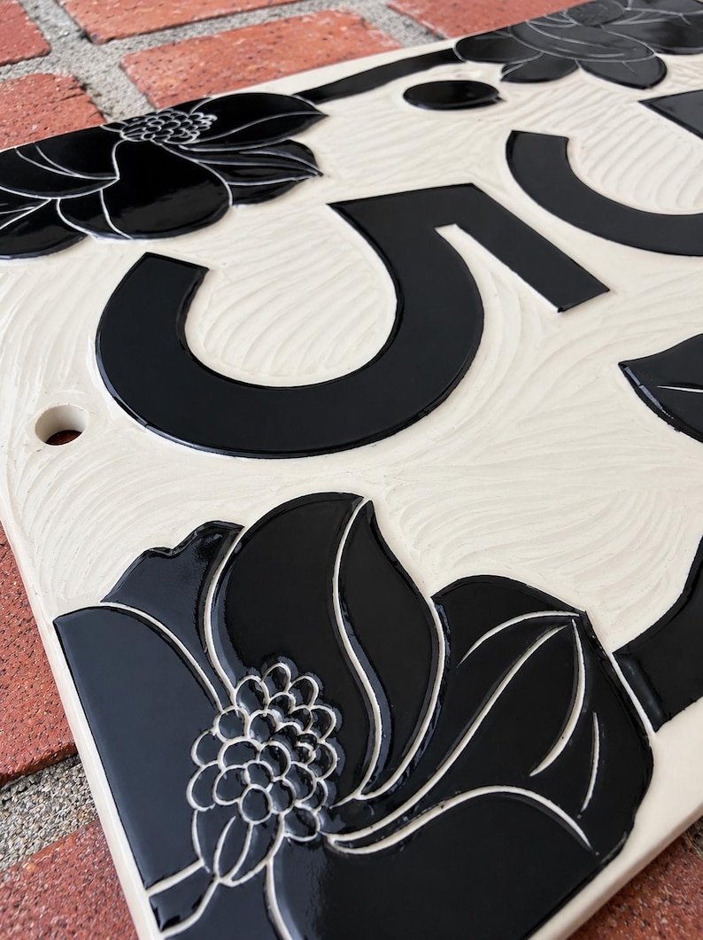 May include: A rectangular ceramic house number plaque featuring a cream-colored background with a textured, wavy surface. The plaque displays a large black numeral five and is decorated with glossy black floral motifs at the corners.