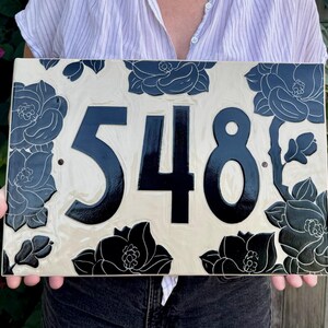 May include: A rectangular ceramic house number plaque featuring the digits 548 in a bold, glossy black font. The background is a creamy off-white color, decorated with intricate, dark black floral illustrations that frame the central numbers.