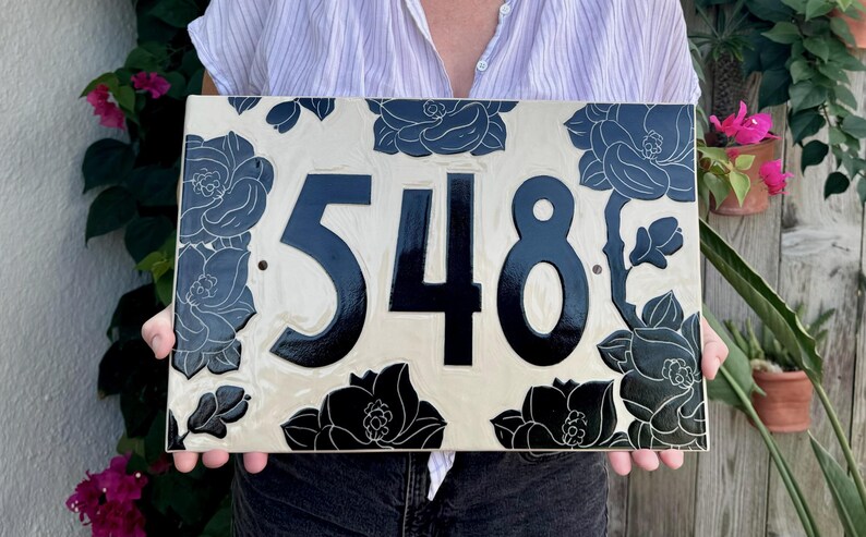 May include: A rectangular ceramic house number plaque featuring the digits 548 in a bold, glossy black font. The background is a creamy off-white color, decorated with intricate, dark black floral illustrations that frame the central numbers.