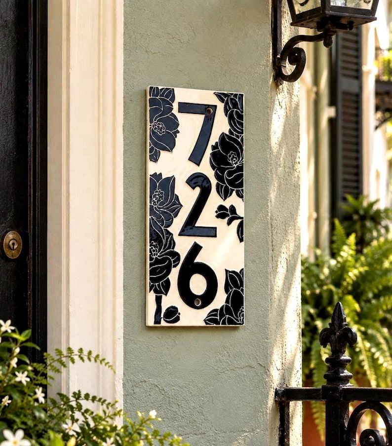 May include: A rectangular house number plaque featuring the digits 726 in a bold black font. The plaque has a cream-colored background decorated with black floral illustrations of flowers and leaves surrounding the centered house numbers.